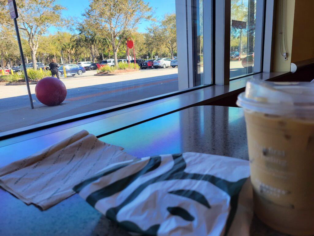 Starbucks coffee and grilled cheese on a  table by a window overlooking cars in the parking lot. 