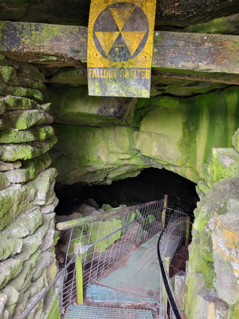 Entrance to the volcanic tube cave at Idaho's Mammoth Cave and Shoshone Bird Museum. The rocks around the entrance are stained green and a sign with a radioactive symbol and "fallout shelter" warning is plastered above the entrance. 